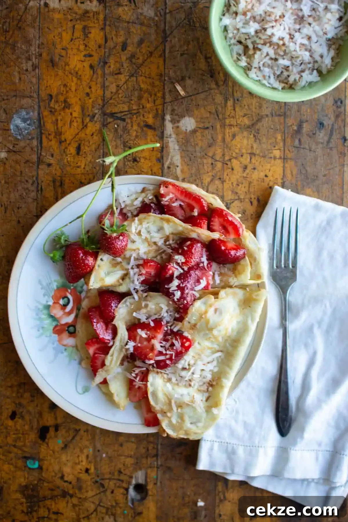 A plate of crepes topped with fresh, sliced strawberries on a wood table with a bowl of toasted coconut behind it. 