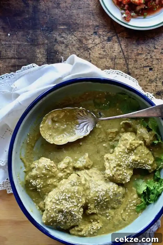 A platter of mole verde chicken with a spoon in the bowl sitting on a wood table on top of a white napkin with a white plate behind it. 