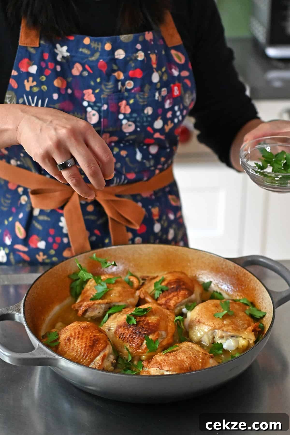 A person in a blue apron is sprinkling fresh parsley leaves onto a skillet filled with lemon garlic chicken.