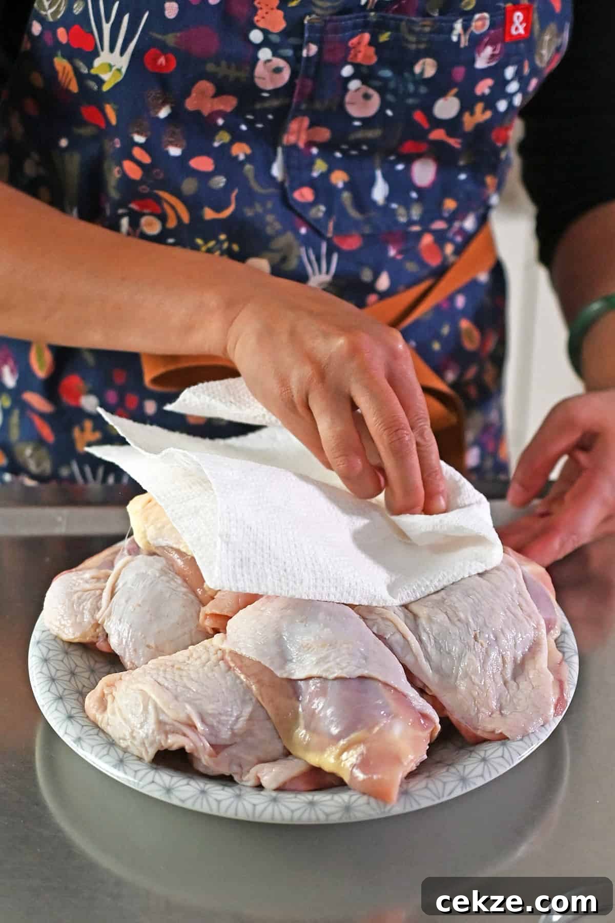 A person in a blue apron is blotting raw skin-on bone-in chicken thighs dry with a paper towel.