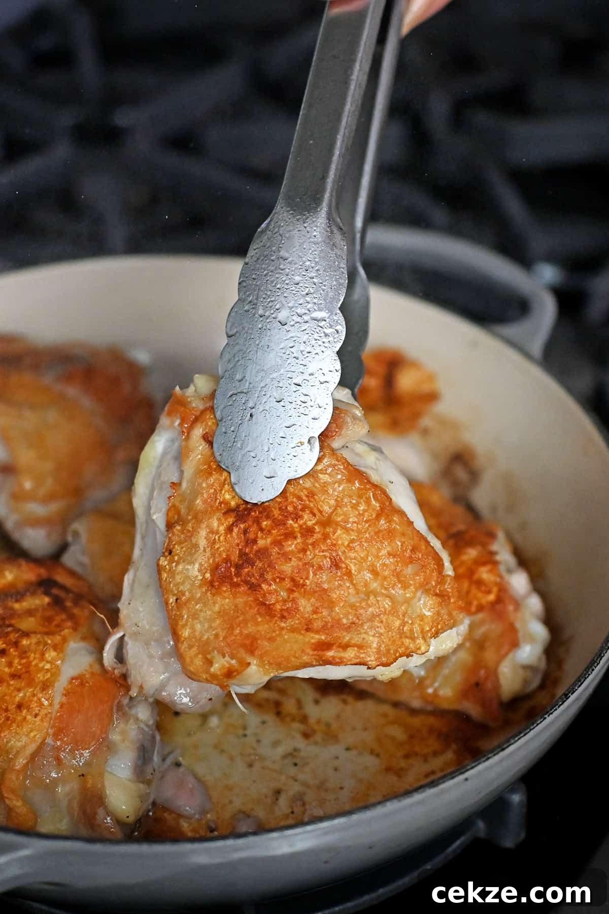 A pair of tongs is picking up a chicken thigh from a skillet with golden brown skin.