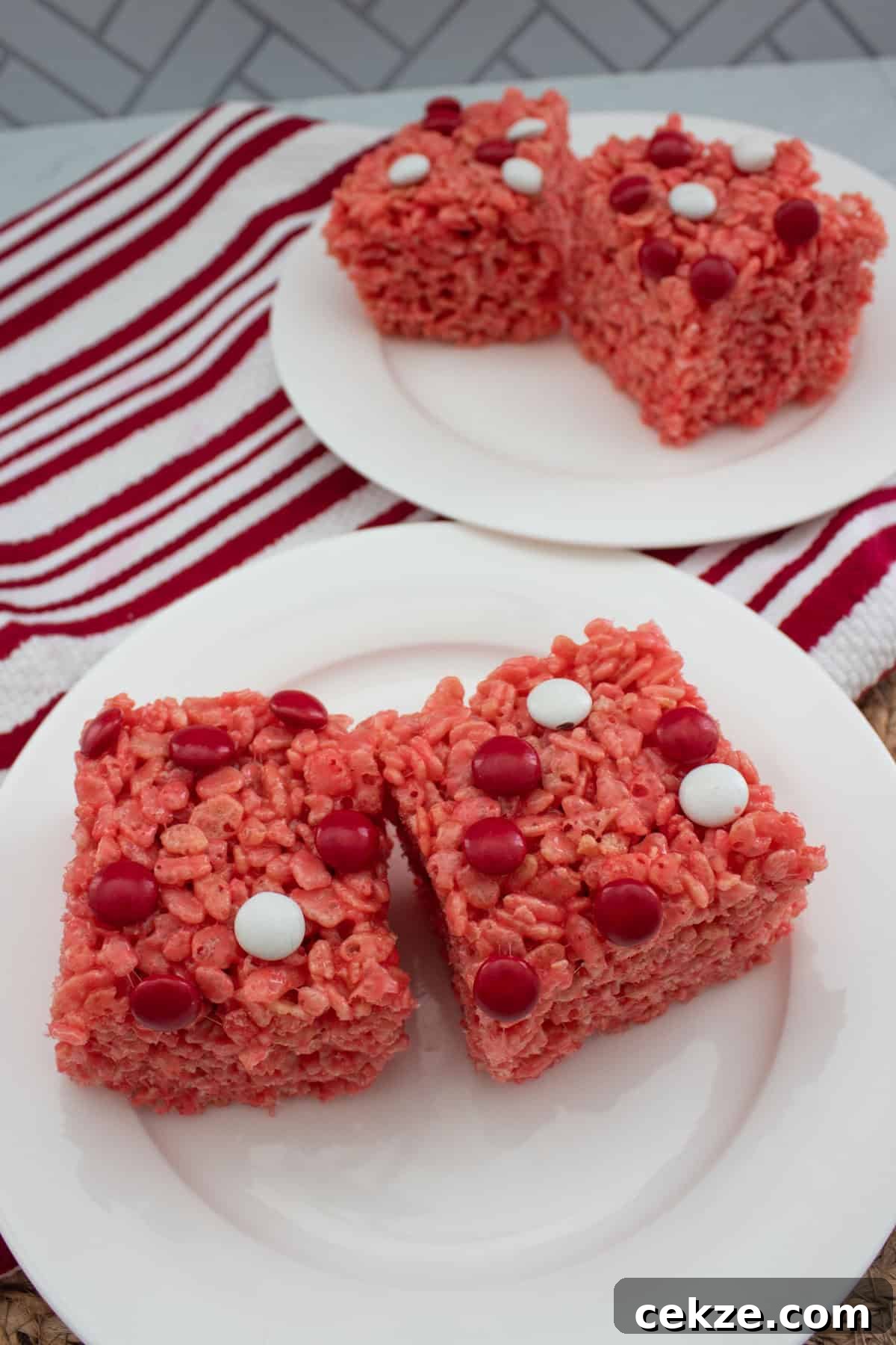 Valentine Rice Krispie Treats: Simple Heart-Shaped Dessert Recipe 2 Two red Rice Krispies treats decorated with red and white candy dots are on a white plate, with more treats on a second plate in the background. A red and white striped cloth is underneath.