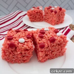 Valentine Rice Krispie Treats: Simple Heart-Shaped Dessert Recipe 11 Two red Rice Krispies treats topped with red and white candy dots are on a white plate in the foreground, with more treats on a second plate in the background. A red and white striped towel is nearby.