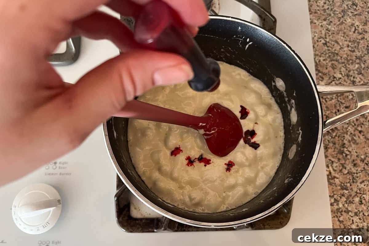 Valentine Rice Krispie Treats: Simple Heart-Shaped Dessert Recipe 5 A hand holds a dropper over a saucepan of thick white mixture on a stove, adding drops of red food dye and stirring with a red spatula.