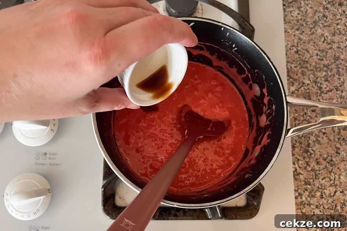 Valentine Rice Krispie Treats: Simple Heart-Shaped Dessert Recipe 6 A hand pours a small cup of vanilla extract into a saucepan with a thick red mixture being stirred on a stove.