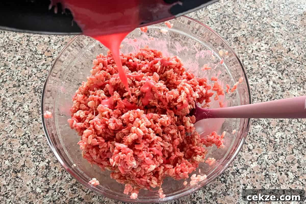 Valentine Rice Krispie Treats: Simple Heart-Shaped Dessert Recipe 7 A bowl of rice cereal is being mixed with a pink liquid, possibly melted marshmallows and coloring, as more of the mixture is poured in from above. A pink spatula rests in the bowl on a speckled countertop.