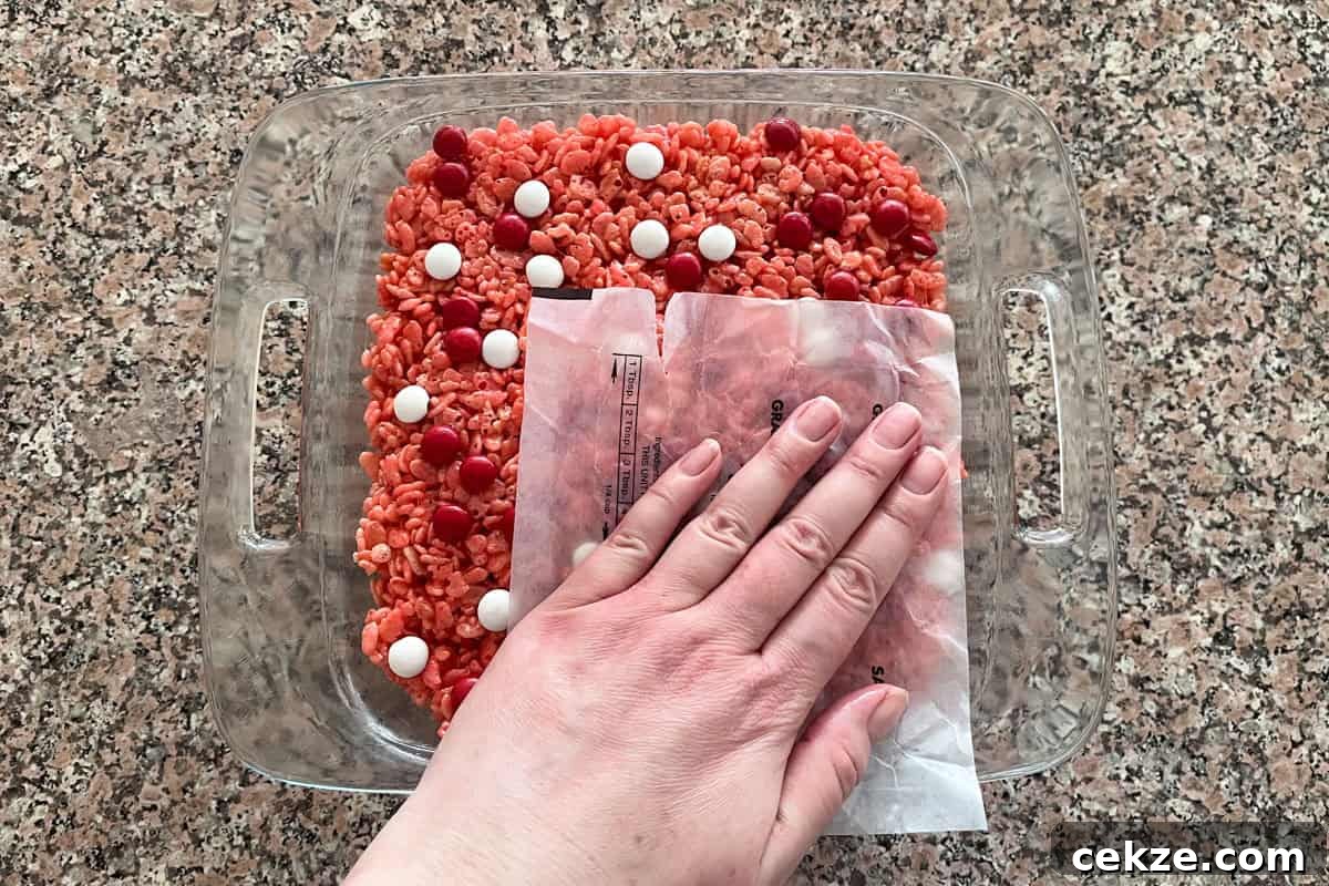 Valentine Rice Krispie Treats: Simple Heart-Shaped Dessert Recipe 8 A hand presses a sheet of wax paper over a layer of red Rice Krispies treat mix with red and white candy pieces in a glass baking dish on a speckled countertop.