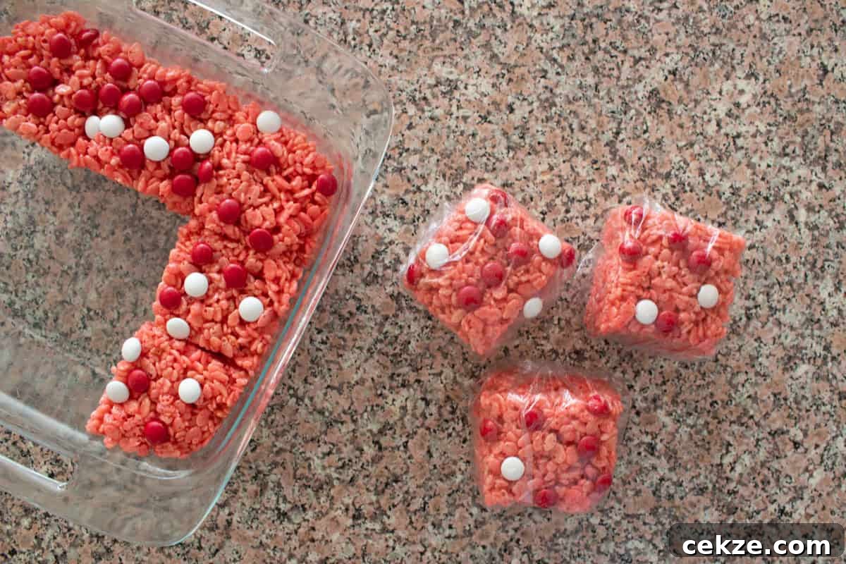 Valentine Rice Krispie Treats: Simple Heart-Shaped Dessert Recipe 9 A glass dish with pink rice crispy treats topped with red and white candies, next to three wrapped individual squares of the same treat, all on a granite countertop.