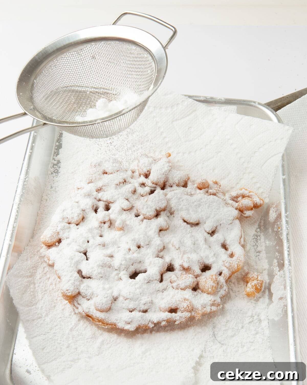 funnel cake on paper towel lined tray being sprinkled with powdered sugar using mesh colander