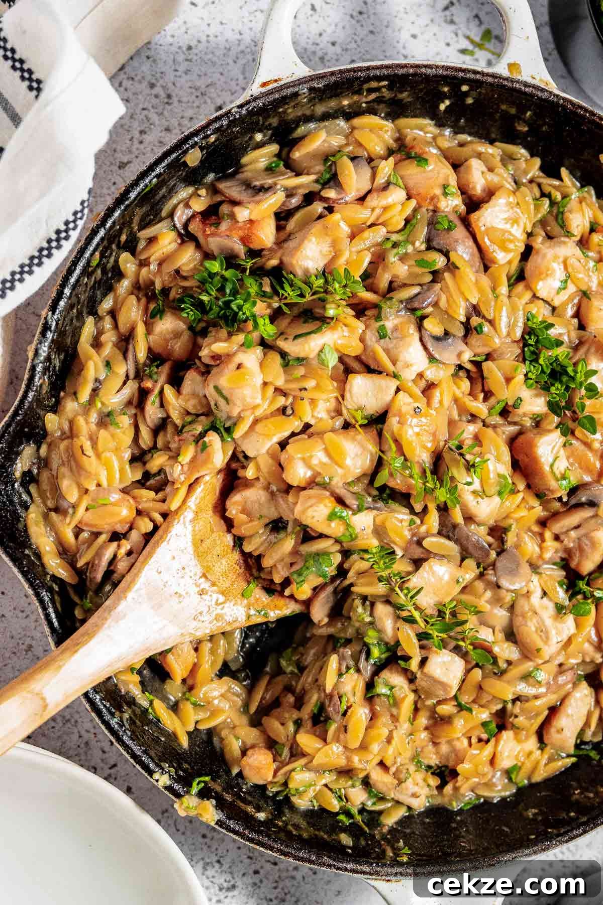 Close up overhead shot of orzo pasta garnished with fresh herbs in a large skillet with a wooden spoon and white cloth on the side.