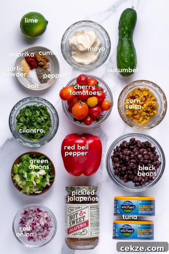 Overhead view of Mexican tuna salad ingredients including albacore tuna, black beans, corn salsa, cherry tomatoes, cucumber, red bell pepper, red onion, green onions, cilantro, lime, pickled jalapeños, mayo, and spices like cumin and paprika.