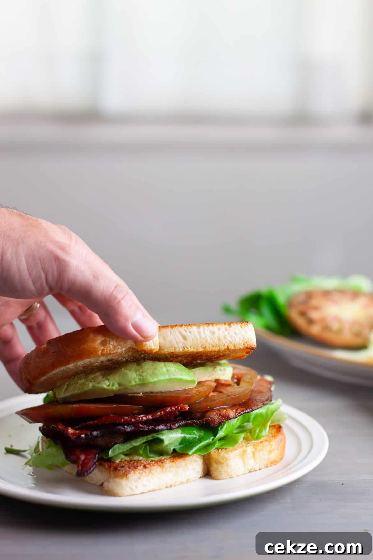 A hand pressing the top slice of bread onto an avocado BLT.