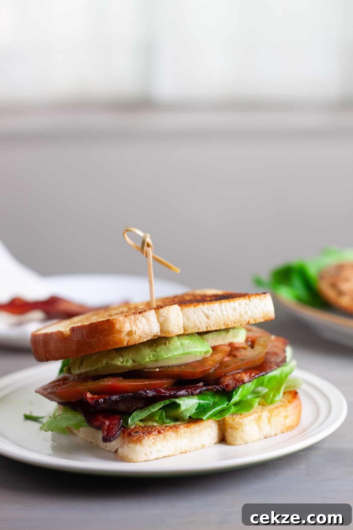 An avocado BLT on a toasted white bread on top of a white plate.