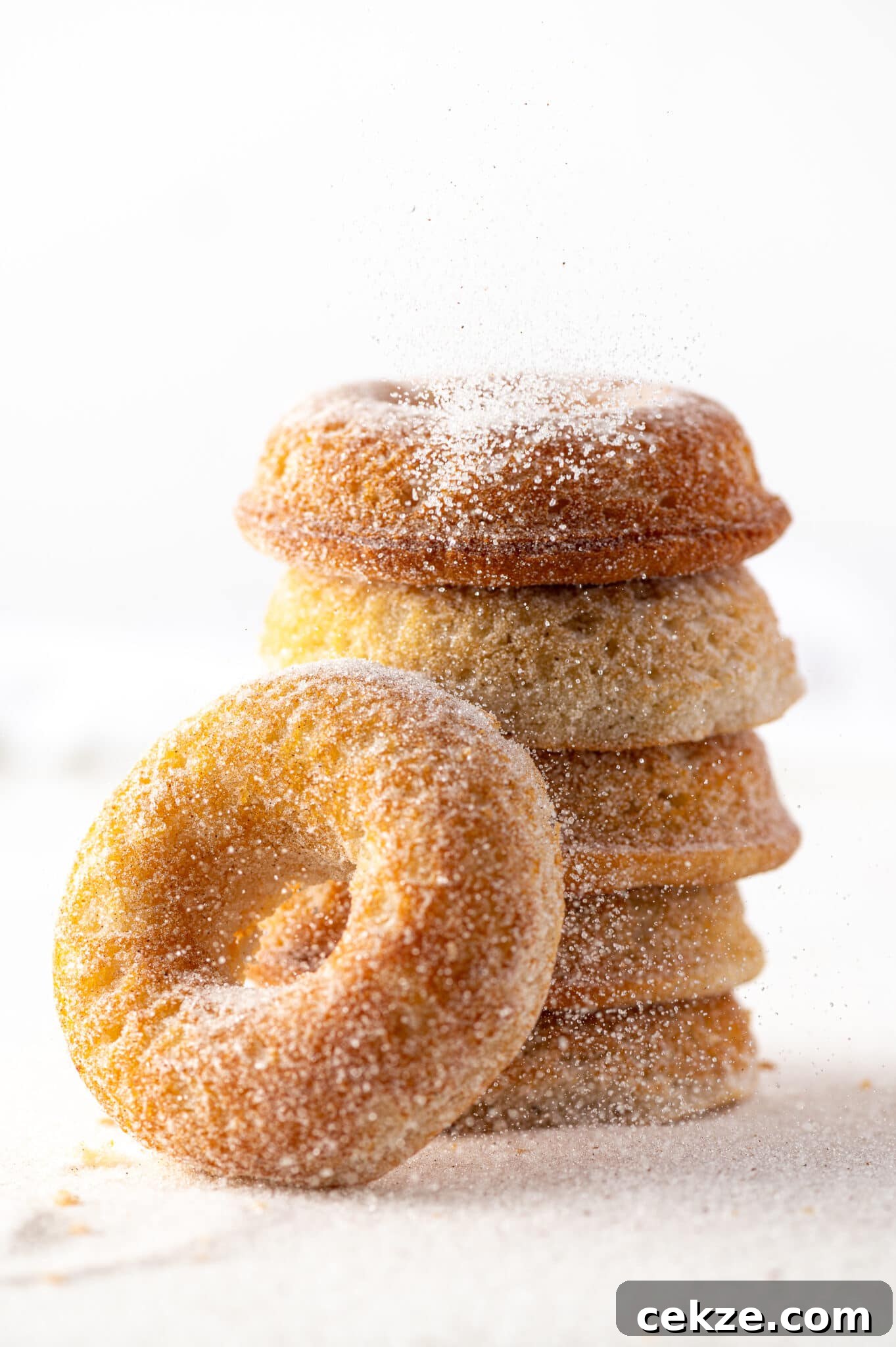 Old fashioned gluten free donuts stacked against a bright white background with cinnamon sugar being sprinkled over the top. 