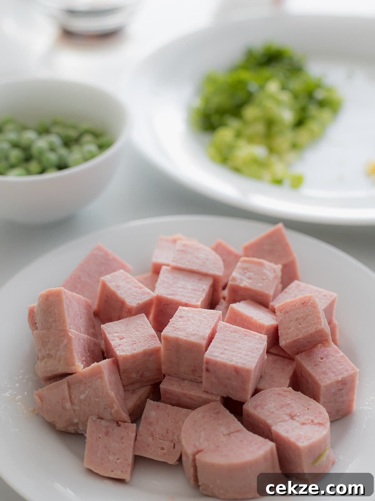 spam cut into cubes on a white plate next to green peas in a bowl