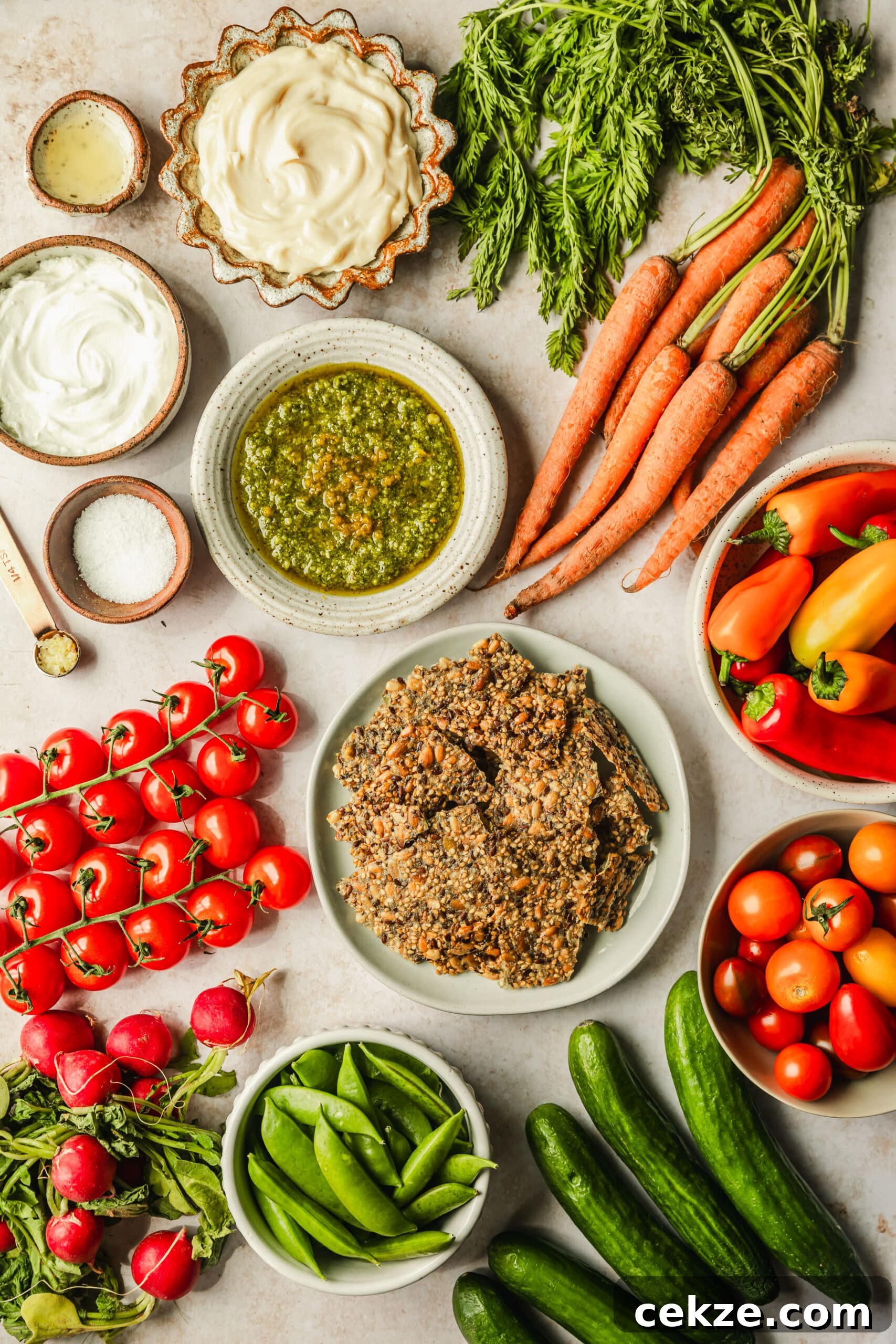White and brown bowls of basil sauce, mayo, yogurt, salt, garlic, lemon juice, crackers, cherry tomatoes, peppers, and peas on a tan counter next to mini cucumbers, radishes, and carrots.