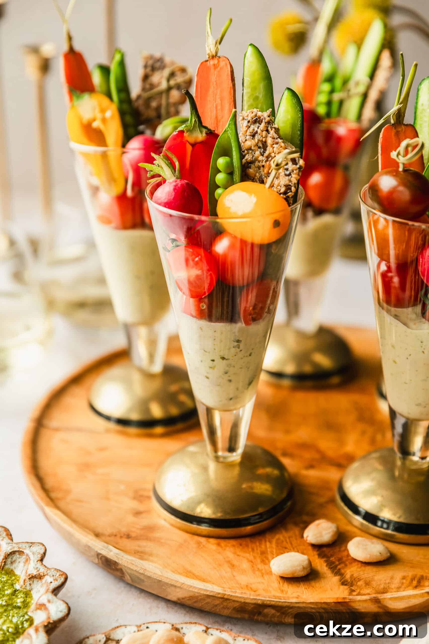 A wood tray of veggie cups with basil sauce on a tan counter next to glasses of white wine, a vase of yellow flowers, and a white plate of almonds.
