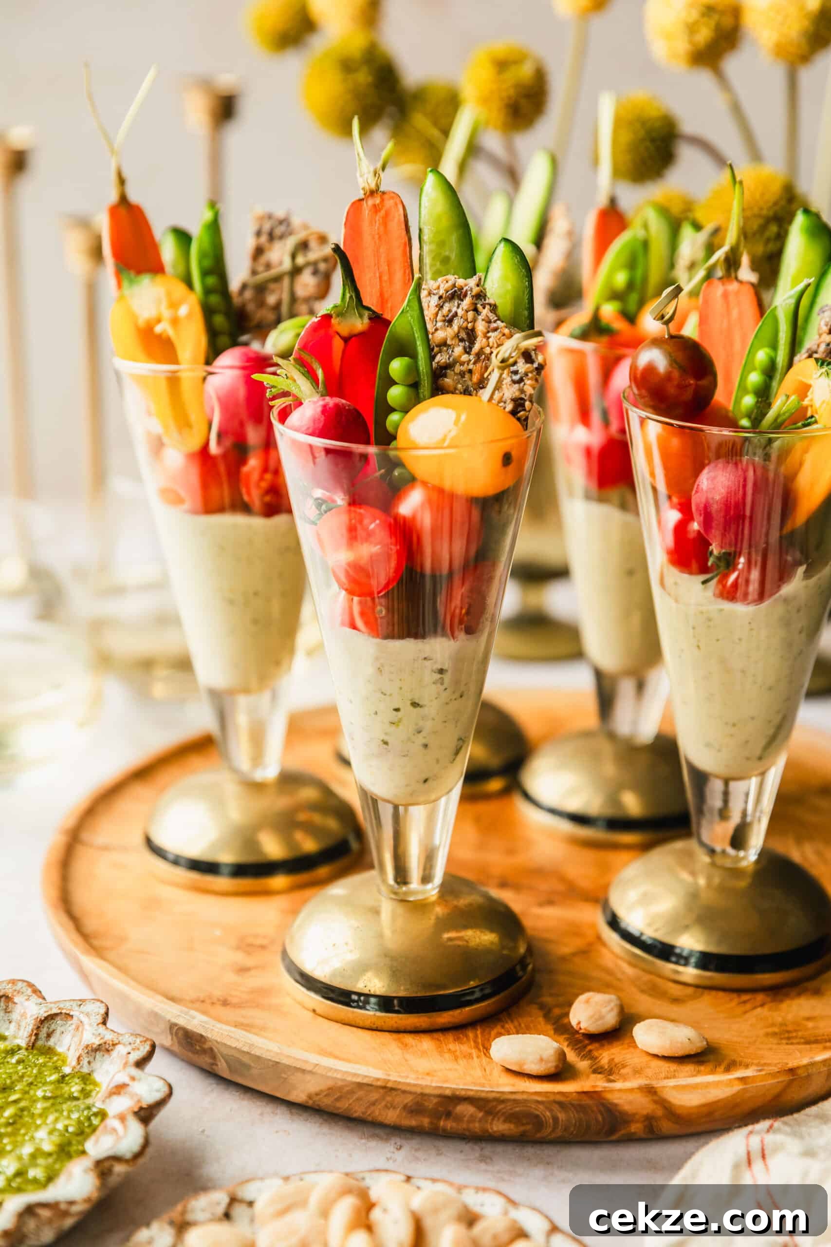 A wood board of crudités cups with pesto dip on a tan counter next to yellow flowers, and white bowls of pesto and almonds.