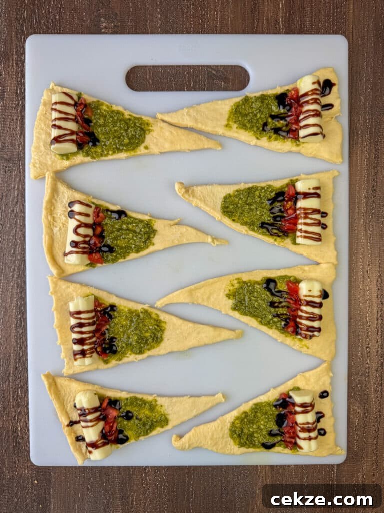 Pesto spread on several unwrapped crescent rolls with mozzarella cheese and a drizzle of balsamic glaze with a chopped cherry tomato on a cutting board.
