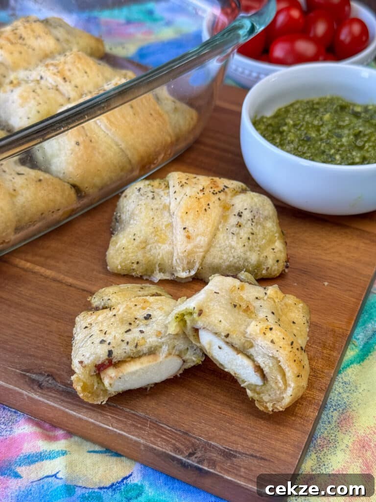 Warm mozzarella cheese filled pesto crescent roll cut in half on a cutting board in front of another roll with pesto and tomatoes in the background.