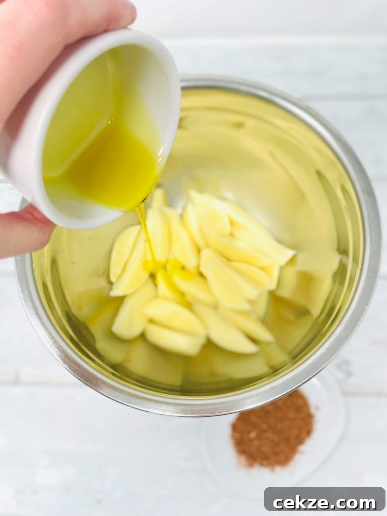 olive oil being poured over freshly cut potato wedges in a stainless steel bowl