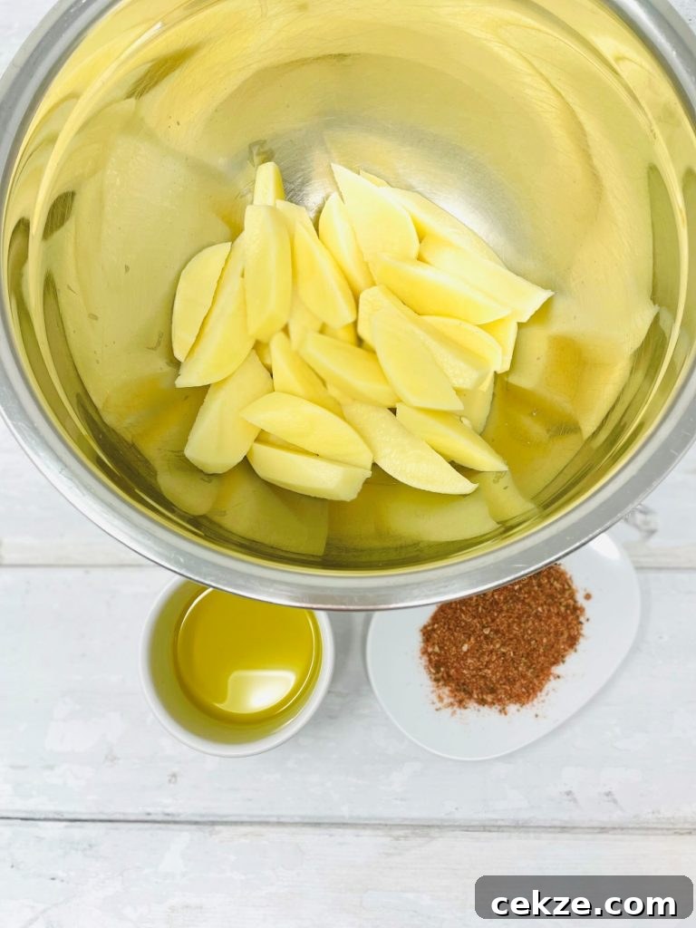 peeled potatoes sliced into wedges in a stainless steel bowl, pot of olive oil and saucer of peri peri seasoning to the side of the bowl