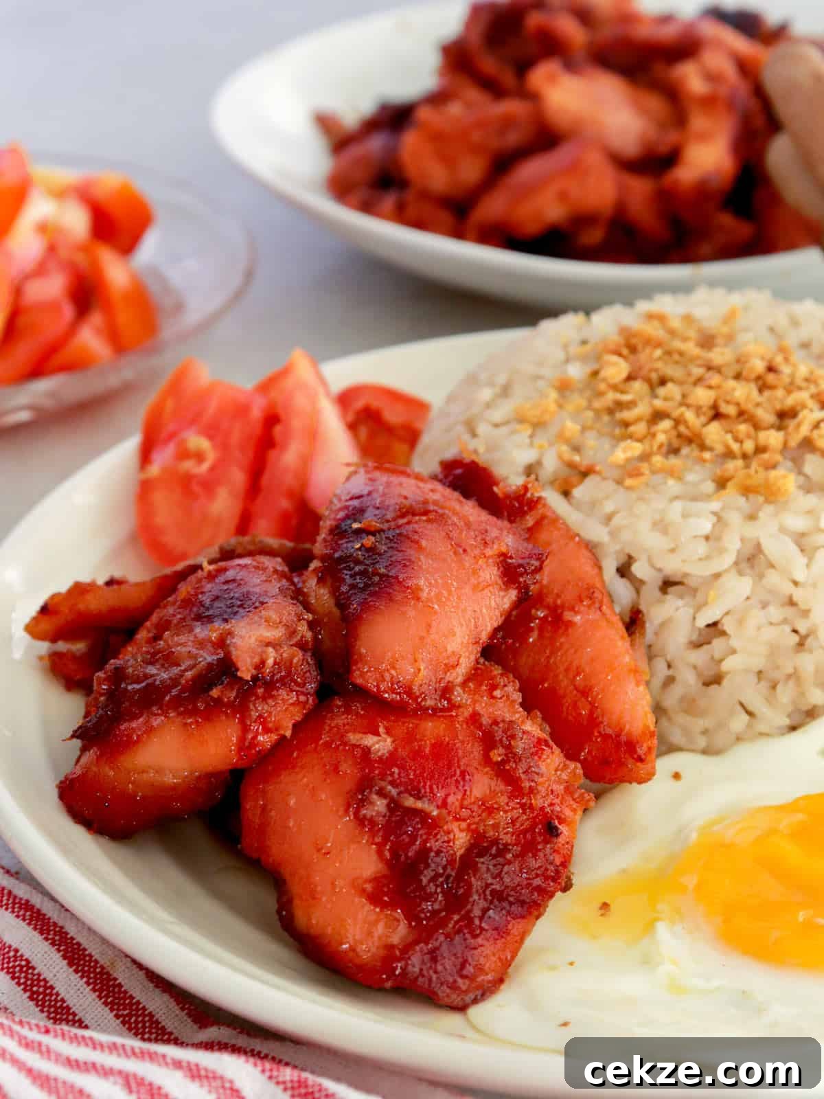 chicken tocino on a white plate with a side of fried egg, garlic rice, and sliced tomatoes.