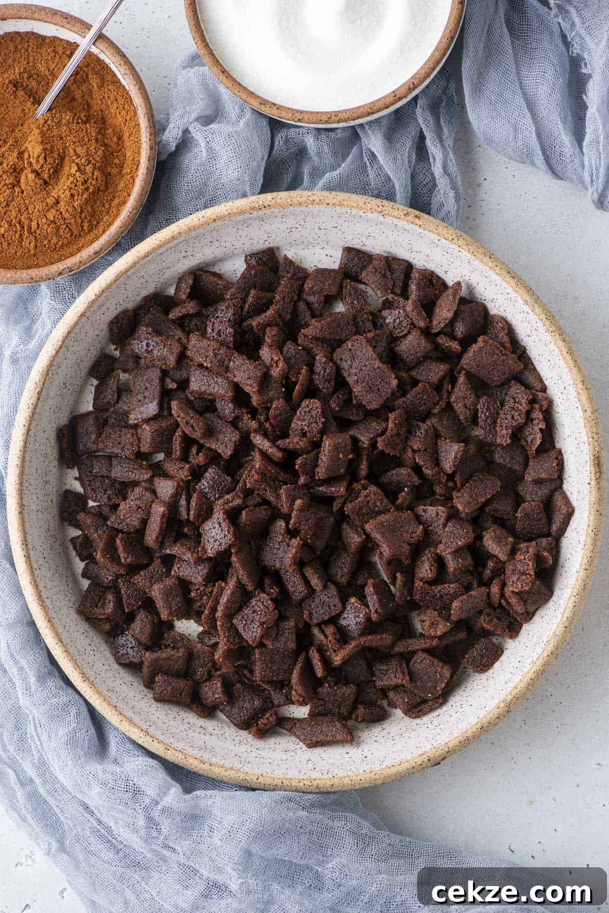 Cinnamon Baking Chips: Flavorful Mix-Ins for Cookies & Muffins 2 A bowl filled with cinnamon chips sits on a light gray surface, with bowls of cinnamon powder and sugar nearby. A blue cloth is draped in the background.