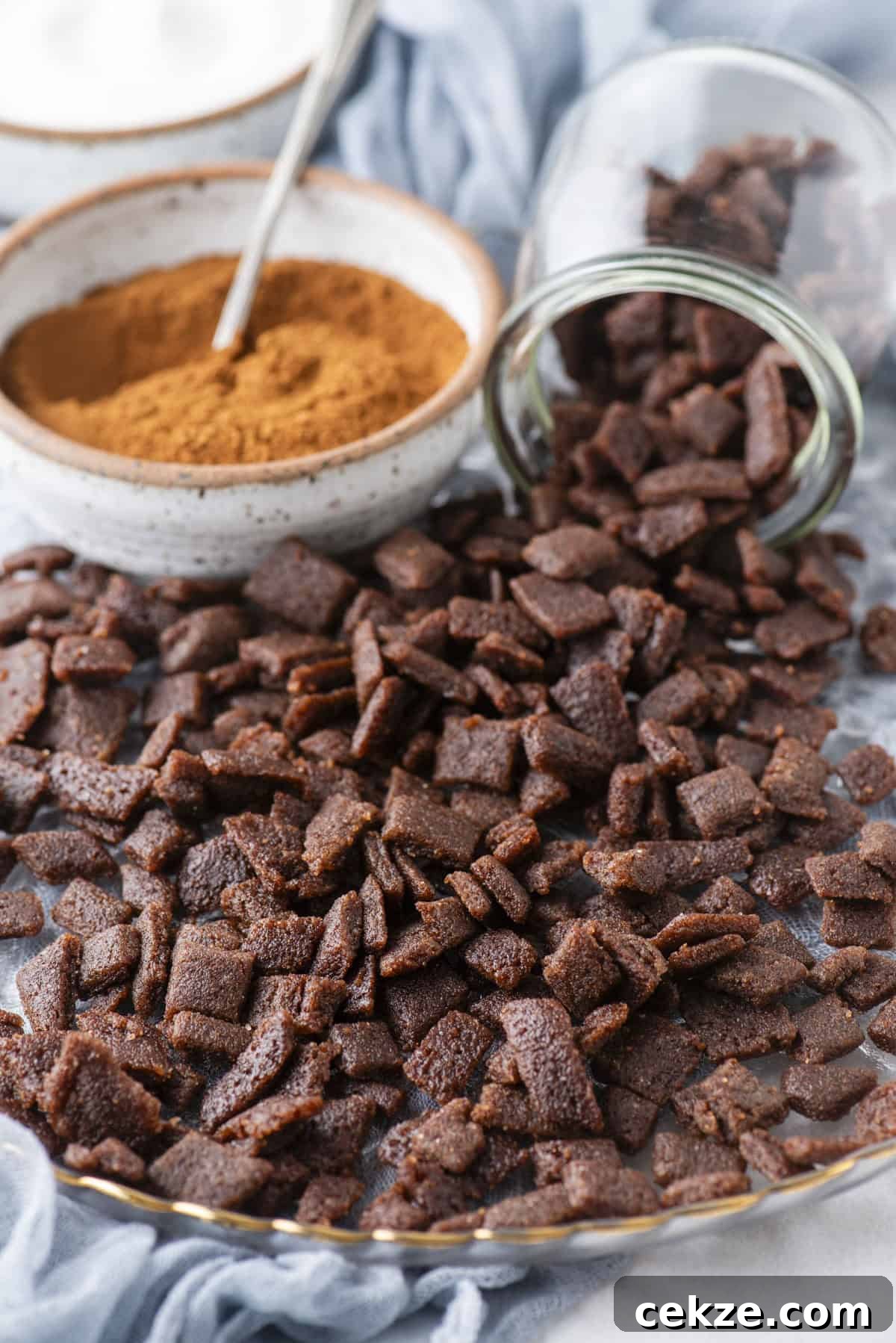 Cinnamon Baking Chips: Flavorful Mix-Ins for Cookies & Muffins 3 A tray covered with cinnamon chips for baking, next to a jar tipped over and a bowl of cinnamon powder with a spoon inside. A light blue cloth is in the background.