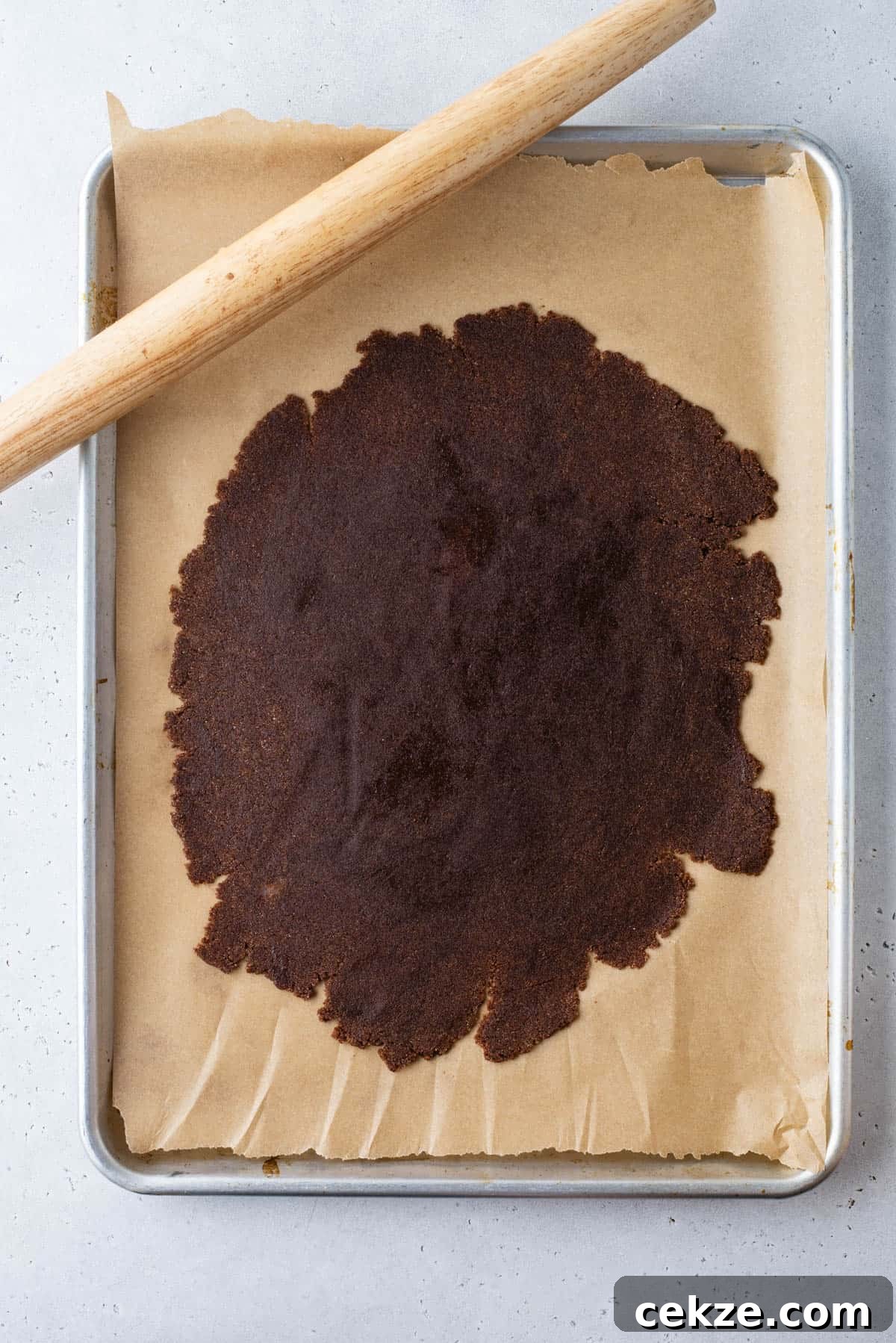 Cinnamon Baking Chips: Flavorful Mix-Ins for Cookies & Muffins 7 A baking tray lined with parchment paper holds a large, thin sheet of dark dough, partially rolled out. A wooden rolling pin rests diagonally along the top edge of the tray.