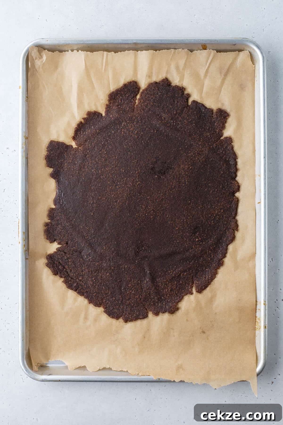 Cinnamon Baking Chips: Flavorful Mix-Ins for Cookies & Muffins 8 A baking sheet lined with parchment paper holds a large, unevenly spread circle of dark brown dough or mixture. The surface is slightly rough in texture, and the edges are irregular.