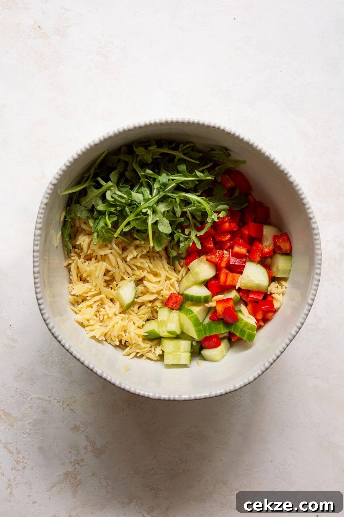 Cooked orzo in a bowl topped with arugula, diced cucumber, and red bell pepper before tossing.