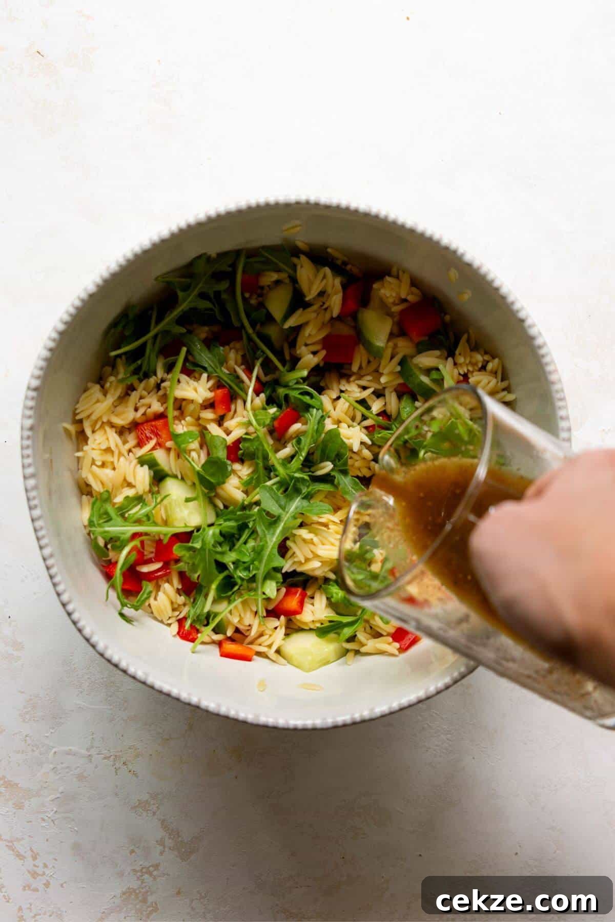 Balsamic dressing being poured over orzo and chopped vegetables in a large bowl.