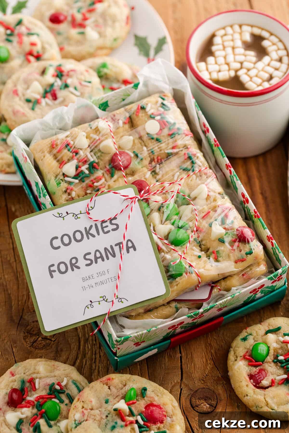 Break-and-Bake Christmas Cookie Dough Tips for Perfect Cookies 2 A holiday gift box filled with rectangular cookie bars topped with red and green M&Ms, white chocolate chips, and sprinkles, wrapped in plastic and tied with red and white string. A label reads “COOKIES FOR SANTA – BAKE 350 F, 11–14 MINUTES.” The box is surrounded by whole cookies, a holly-decorated plate, and a white mug of hot cocoa topped with mini marshmallows.