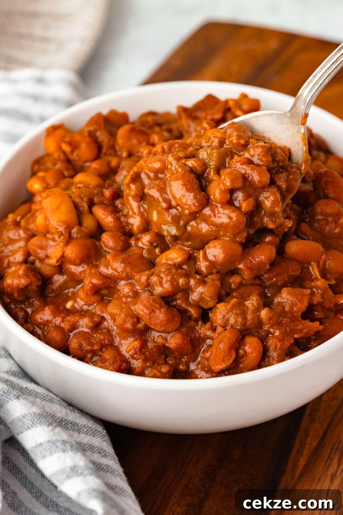 A white bowl filled with baked beans in thick sauce, with a spoon scooping some out. The bowl is placed on a wooden surface next to a striped cloth.