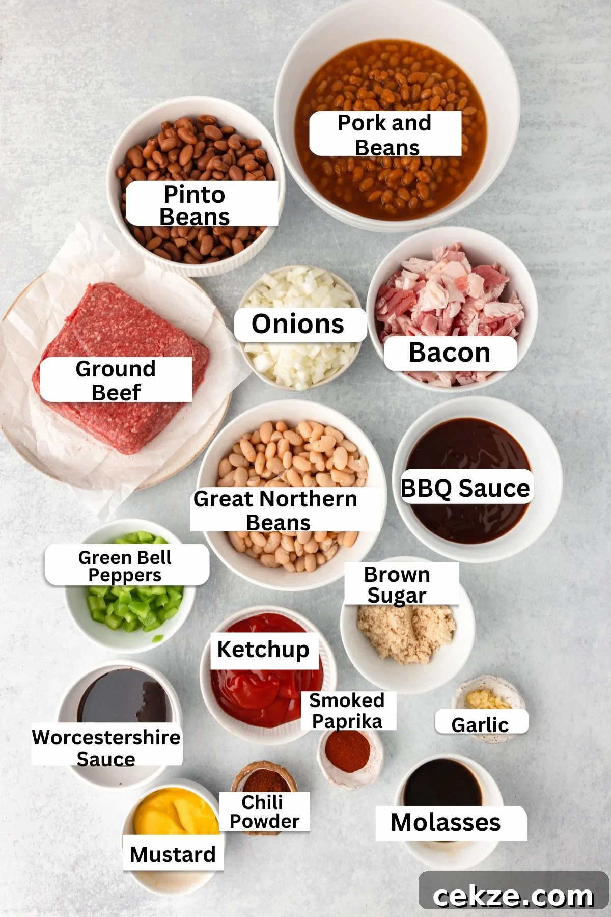 An assortment of ingredients for Cowboy Baked Beans in bowls and containers includes pork and beans, bacon, onions, ground beef, pinto and great northern beans, BBQ sauce, brown sugar, spices, ketchup, green bell peppers, mustard, Worcestershire sauce, and molasses.