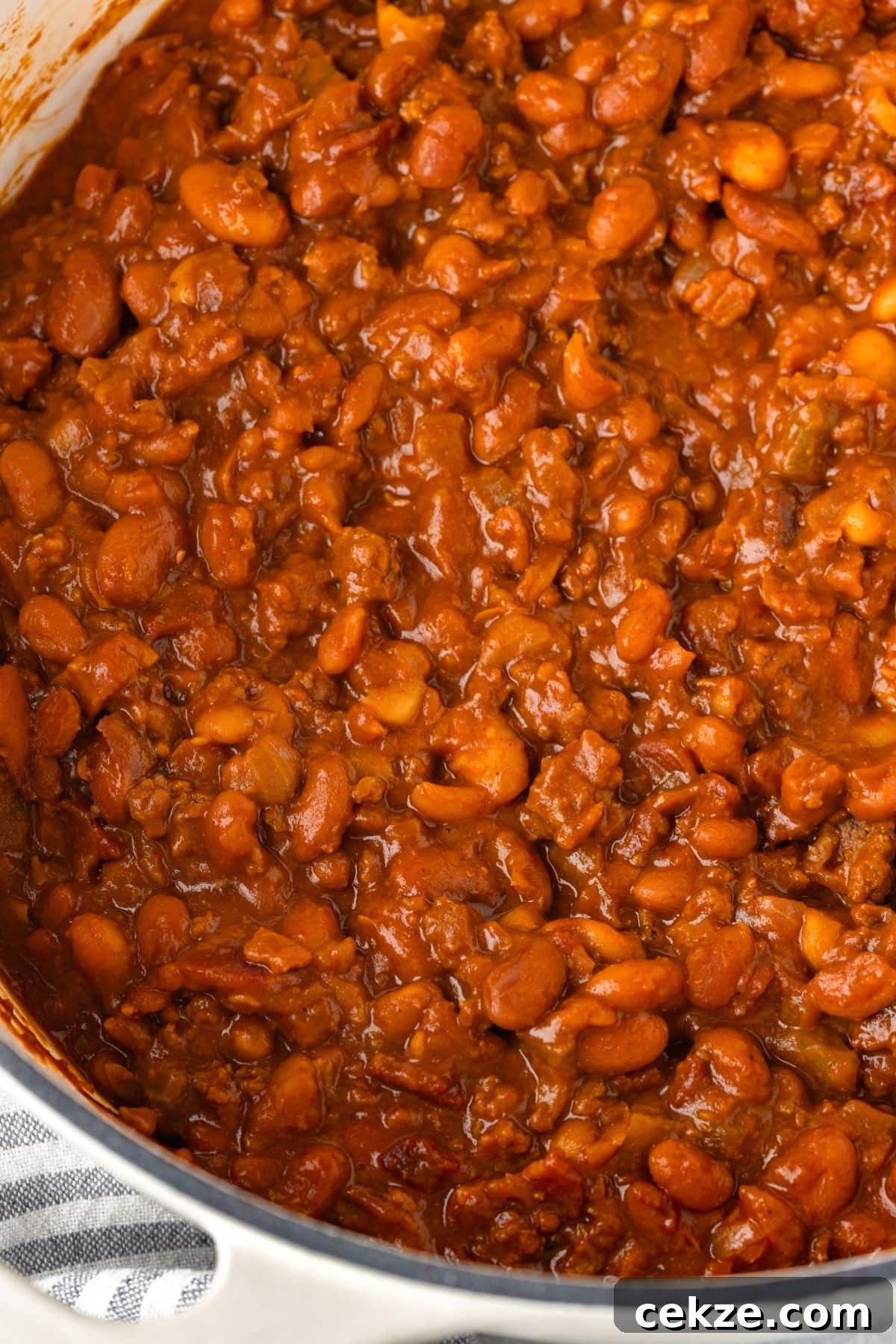 Close-up of a pot filled with baked beans in a rich, tomato-based sauce. The beans are plump and coated with the sauce, showing a hearty, savory dish. A small part of the pots rim is visible on the bottom right.
