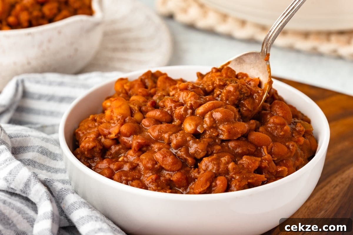 A white bowl filled with baked beans in a thick, rich sauce, with a spoon resting inside. The bowl is placed on a wooden surface, and a striped cloth napkin lies beside it. A blurred bowl is in the background.