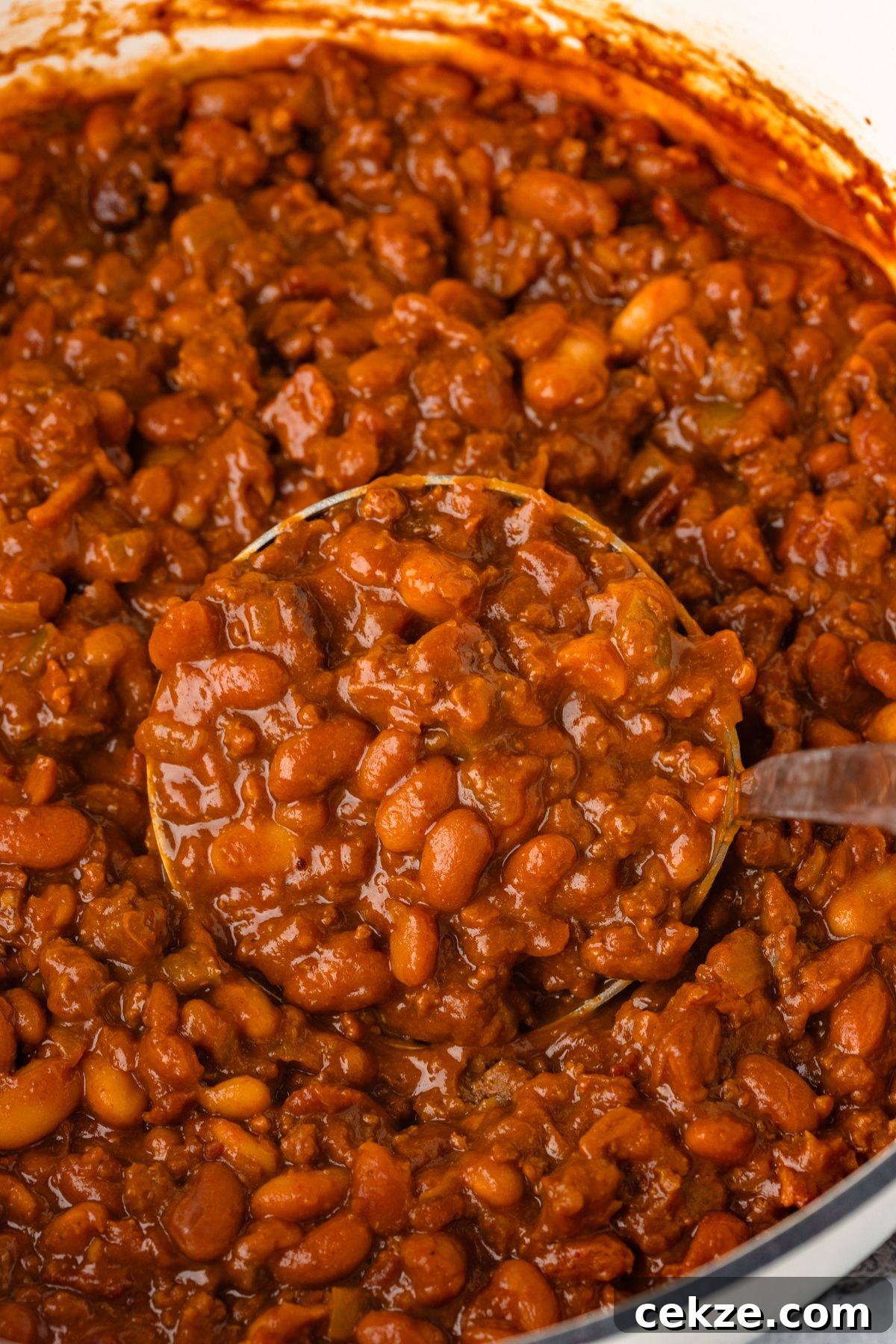 Close-up of a pot filled with savory baked beans in a rich, reddish-brown sauce. A ladle is scooping up the beans, showing their thick texture and hearty appearance.