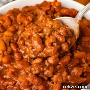 A close-up of Cowboy Baked Beans in a thick, rich sauce, with a spoon lifting a portion from a white bowl.