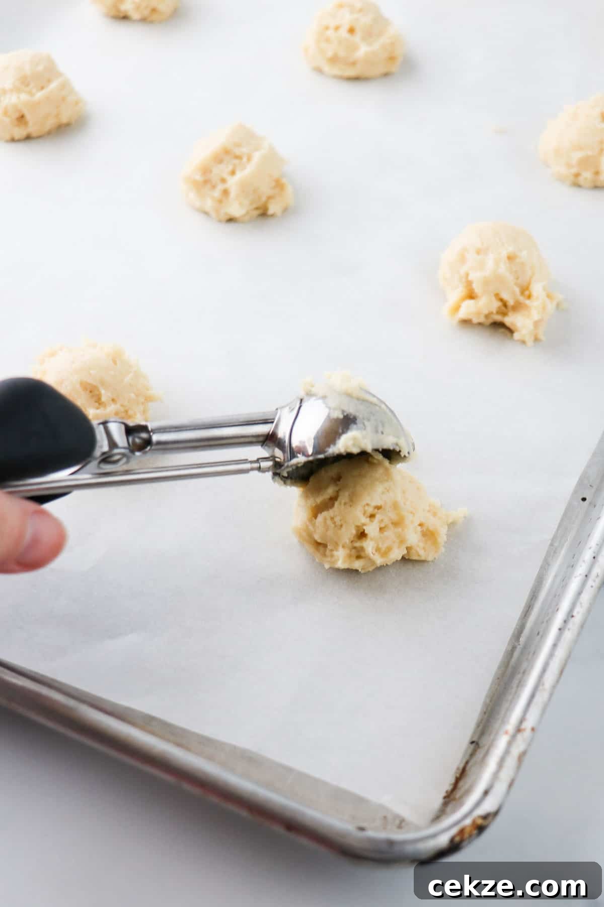 Person scooping Old-Fashioned Buttermilk cookie dough onto a baking sheet with metal scoop.