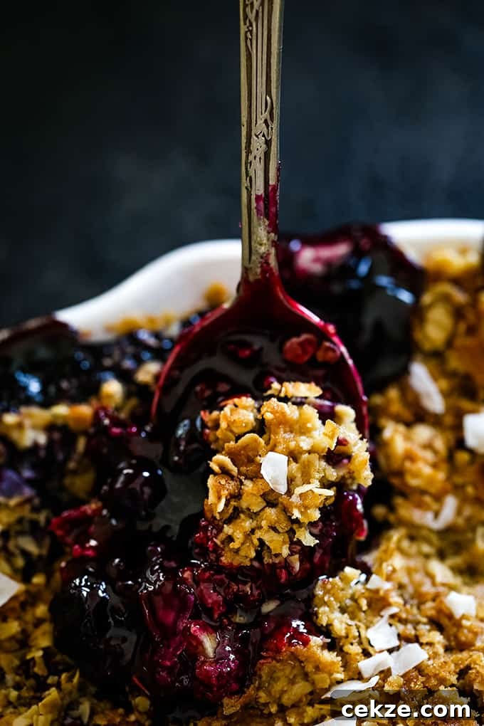 It takes just 15 minutes to prepare this delicious Coconut Blueberry Crisp featuring warm, bubbly blueberries capped by a crunchy cap of oats and coconut. A spoon removes a serving of Coconut Blueberry Crisp from a white baking dish.
