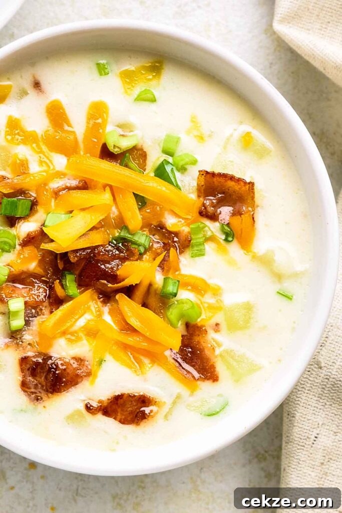 Bowl of loaded potato soup filled with chunks of potato, melted cheese, green onions, and bacon crumbles on a light background.