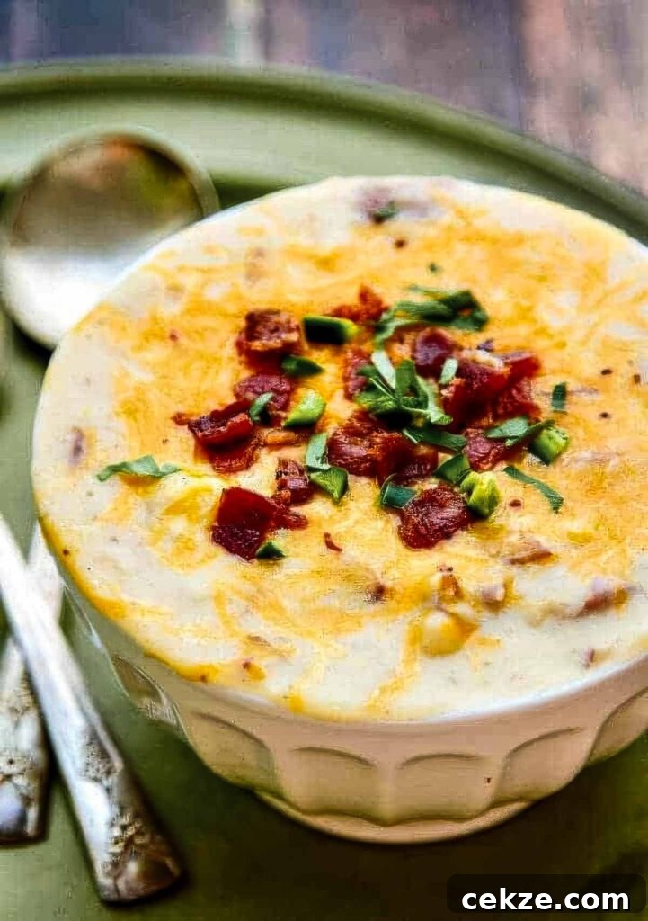 Bowl of thick and creamy potato soup topped with melted cheese, crispy bacon pieces, and fresh herbs on a rustic green plate.