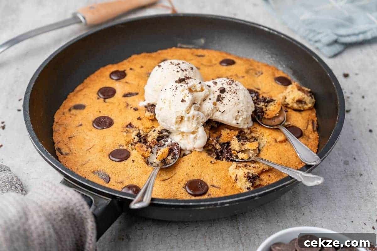 Overhead shot of a warm chocolate chip skillet cookie in a black skillet, topped with scoops of vanilla ice cream and three spoons.