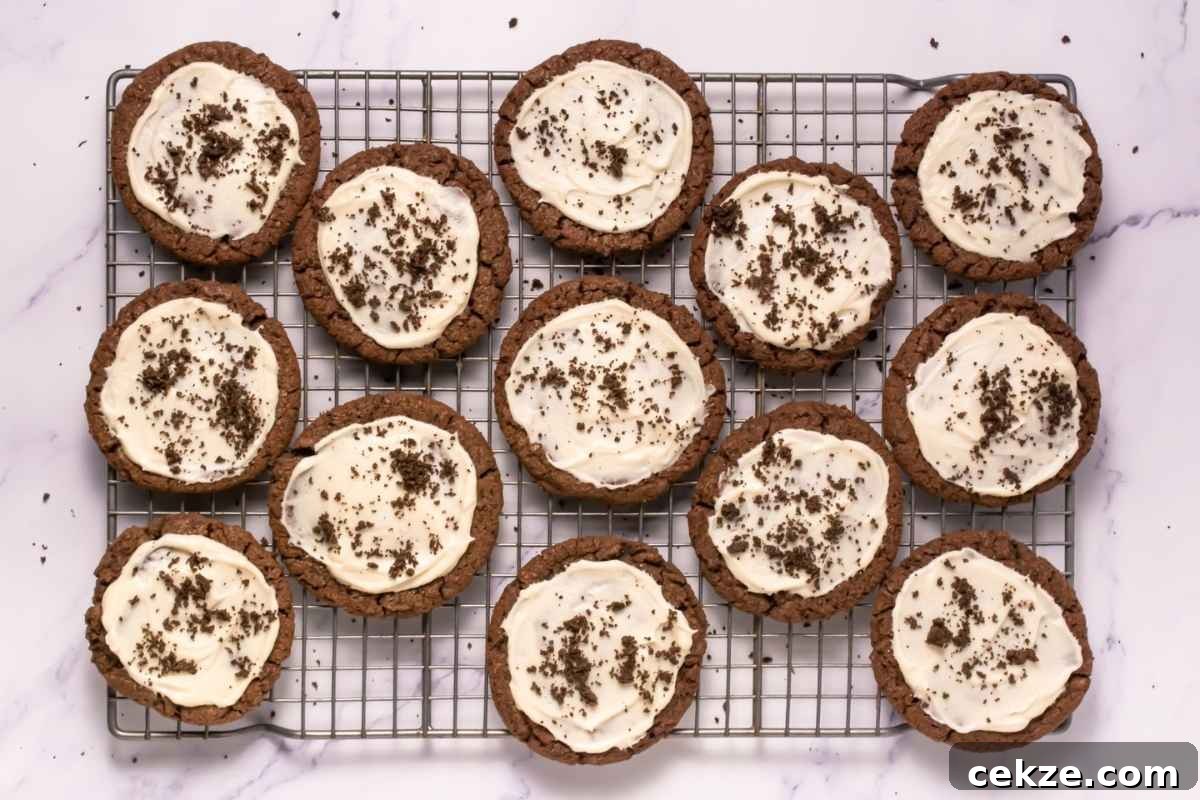A cooling rack with twelve Crumbl Copycat Frosted Oreo Cookies topped with white frosting and chocolate crumbs, arranged on a white marble surface.