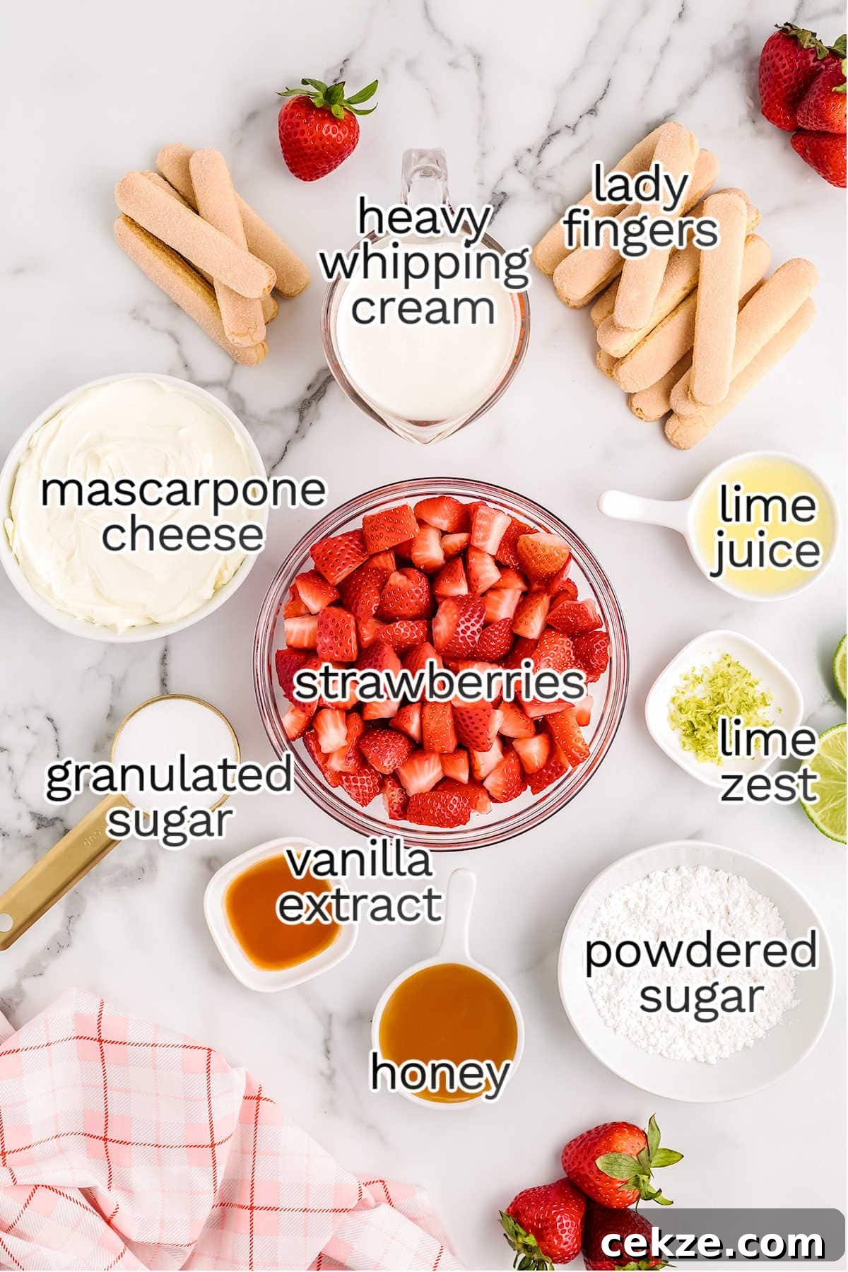 A top-down view of ingredients for a strawberry tiramisu dessert, including strawberries, ladyfingers, mascarpone cheese, heavy whipping cream, granulated and powdered sugar, honey, lime juice, lime zest, and vanilla extract.