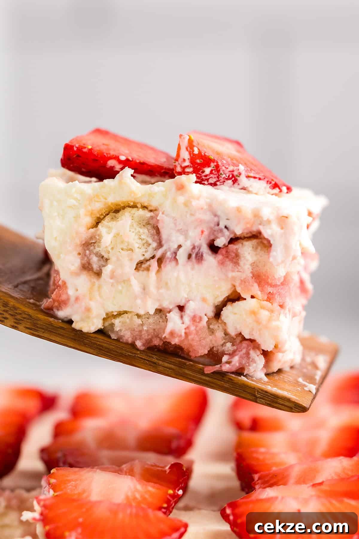 A close-up of a slice of strawberry tiramisu dessert with creamy layers and sliced strawberries on top, served on a wooden spatula.