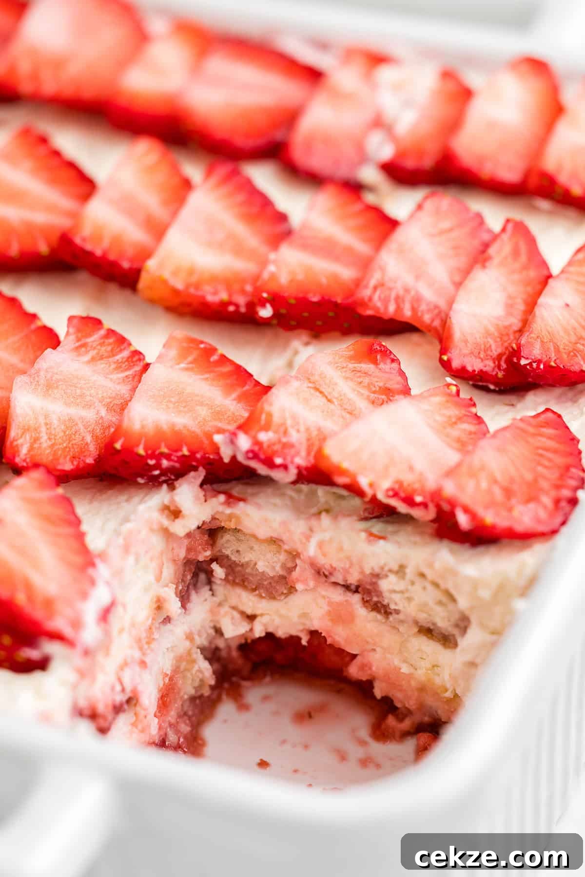 A close-up of a partially eaten strawberry tiramisu in a white dish, topped with sliced strawberries and cream, with delicious layers visible underneath.