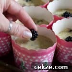 Woman placing a fresh blackberry on top of a muffin case filled with blackberry muffin batter